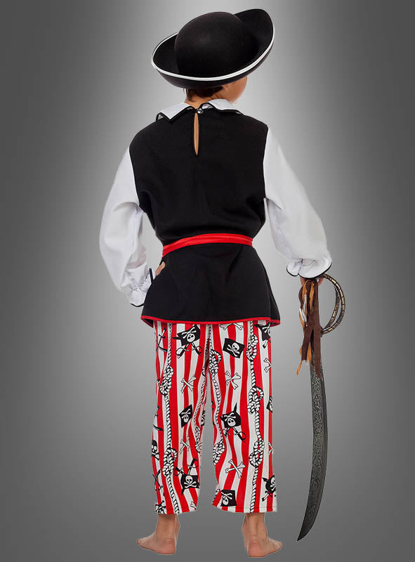 Ein Kind im Piratenkostüm „Captain Seadog Kinder“, das einen schwarzen Hut, ein weißes Hemd, eine schwarze Weste, eine rote Schärpe und gestreifte Hosen mit Totenkopfmuster trägt, hält ein Spielzeugschwert in der Hand und steht barfuß mit dem Rücken zur Kamera.
