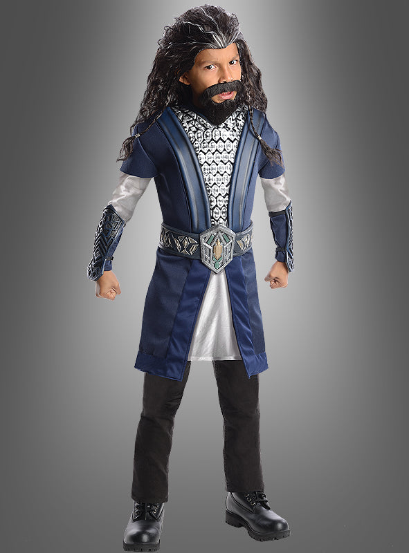 Ein Kind mit einer langen, dunklen Perücke und einem Bart trägt das Thorin-Kinderkostüm aus „Der Hobbit“ in Blau, Silber und Schwarz und steht mit geballten Fäusten vor einem grauen Hintergrund.