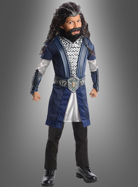 Ein Kind mit einer langen, dunklen Perücke und einem Bart trägt das Thorin-Kinderkostüm aus „Der Hobbit“ in Blau, Silber und Schwarz und steht mit geballten Fäusten vor einem grauen Hintergrund.