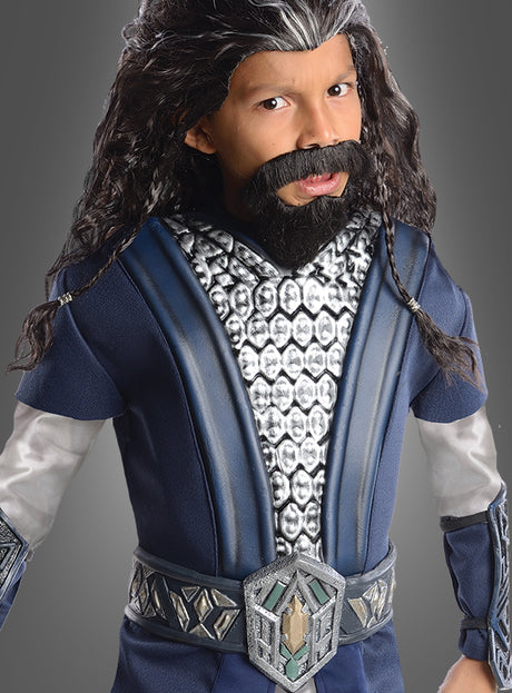 Ein Kind im Thorin-Kinderkostüm aus „Der Hobbit“ – mit langer schwarzer Perücke, Kunstbart, blau-silberner Tunika und Ziergürtel – posiert vor einem grauen Hintergrund – perfekt für junge Fans des offiziellen „Der Hobbit“-Lizenzkostüms.