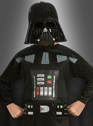 Ein Kind, das das Darth-Vader-Kinderkostüm aus der Kollektion der offiziellen Lizenzkostüme trägt – bestehend aus einem schwarzen Helm, einer Maske, einem Umhang und einem Brustpanzer –, steht mit den Händen in die Hüften gestemmt vor einem grauen Hintergrund.