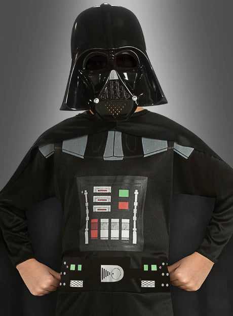 Ein Kind, das das Darth-Vader-Kinderkostüm aus der Kollektion der offiziellen Lizenzkostüme trägt – bestehend aus einem schwarzen Helm, einer Maske, einem Umhang und einem Brustpanzer –, steht mit den Händen in die Hüften gestemmt vor einem grauen Hintergrund.