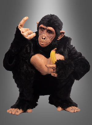 Eine Person, die das „Deluxe Affenkostüm Schimpanse Komplettkostüm“ trägt, hockt mit einer Banane in der Hand, wobei eine Hand erhoben ist und die Finger eine Geste bilden, vor einem grau verlaufenden Hintergrund.