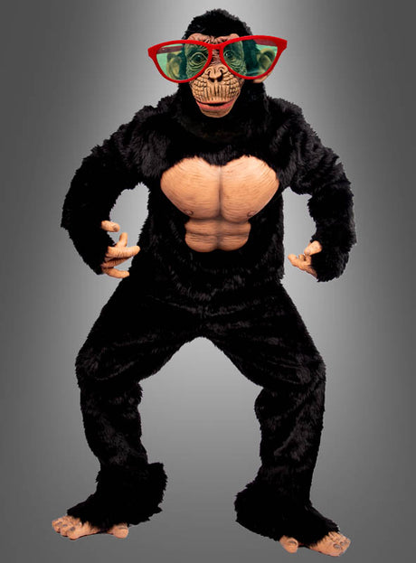 Eine Person, die das „Deluxe Affenkostüm Schimpanse Komplettkostüm“ trägt – mit übertrieben ausgeprägten Bauchmuskeln und einer großen, rot gerahmten grünen Sonnenbrille –, steht vor einem grauen Hintergrund.