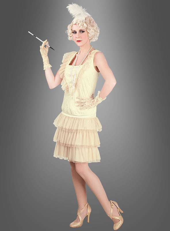 Eine Frau im „Charleston Lady“-Damenkostüm – bestehend aus einem cremefarbenen Flapper-Kleid im Vintage-Stil mit Handschuhen, High Heels und einem Federkopfschmuck – posiert mit einem Zigarettenspitzenhalter vor einem schlichten grauen Hintergrund und verkörpert damit ein elegantes Kostüm im Stil der 20er Jahre.