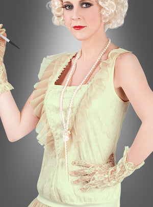 Eine Frau, die das „Charleston Lady“-Damenkostüm trägt – ein leichtes Spitzenkleid, das von Flapper-Kleidern inspiriert ist –, kombiniert es elegant mit Perlen und Spitzenhandschuhen und hält dabei eine Zigarettenspitze in der Hand; ihr kurzes, lockiges blondes Haar und ihr ausdrucksloser Gesichtsausdruck vor einem grau verlaufenden Hintergrund vermitteln das raffinierte Flair des klassischen Kostümstils der 20er Jahre.