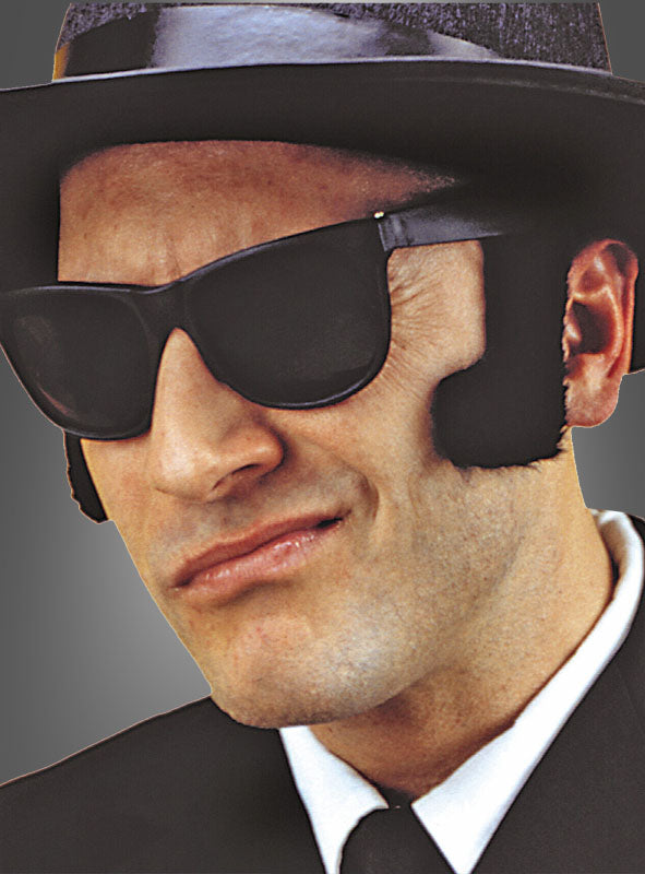 Ein Mann mit schwarzer Sonnenbrille, schwarzem Hut und Anzug, dazu ein weißes Hemd und eine schwarze Krawatte; als Kostümzubehör trägt er Klebe-Koteletten und hat ein leichtes Grinsen auf den Lippen.