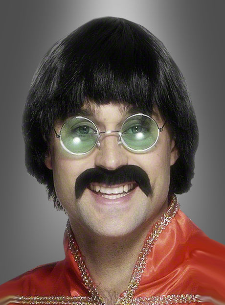 Eine Person, die eine 70er-Beat-Perücke und einen Bart trägt, ergänzt durch eine runde Brille und ein leuchtend rotes Hemd mit goldenem Besatz, lächelt vor einem grauen Hintergrund mit Farbverlauf in die Kamera.