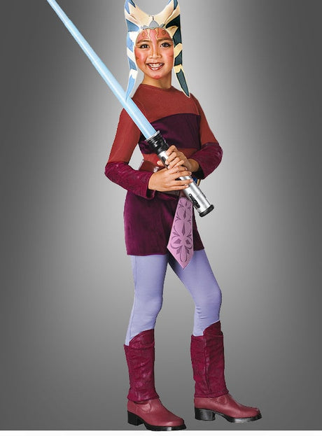 Ein Kind, das ein Ahsoka-Jedi-Kostüm aus „Clone Wars“ von „Star Wars“ trägt und ein blaues Lichtschwert in der Hand hält, steht vor einem grauen Hintergrund und lächelt.