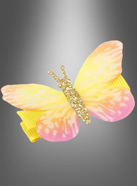 Eine Haarspange „Schmetterling mit Glitzer“ mit einem gelb-rosa Schmetterling und goldenen Glitzerdetails vor einem grauen Hintergrund mit Farbverlauf ist ein bezauberndes Stück Schmetterling-Haarschmuck für jeden Anlass.