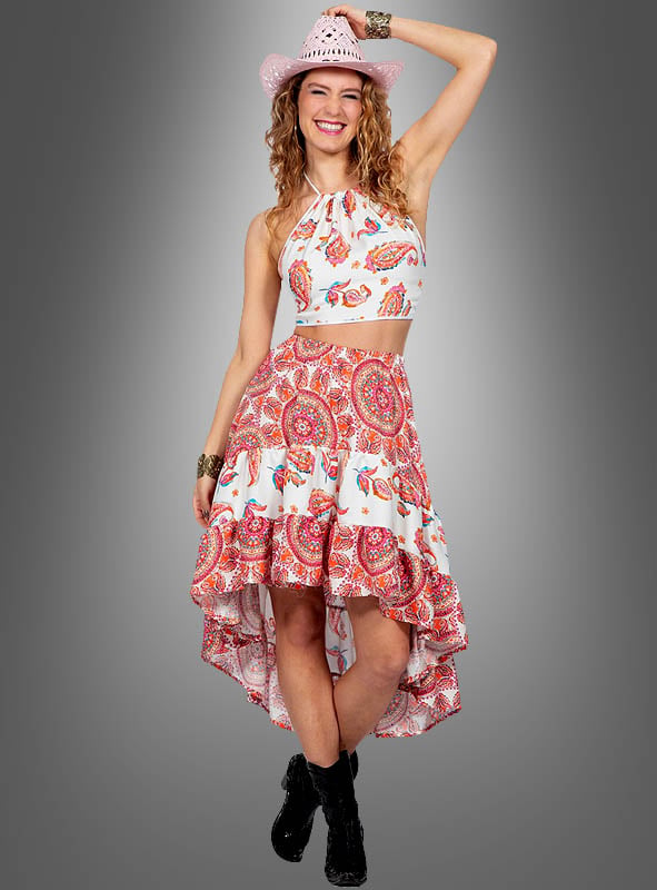 Sie trägt den „Paisley 2-Teiler mit Rock im Festival-Style“ – ein schickes zweiteiliges Set bestehend aus einem Crop-Top mit Paisley-Muster und einem Asymmetrie-Rock – und rundet ihren Festival-Look mit einem rosa Cowboyhut und schwarzen Stiefeln ab, während sie lächelnd vor einem grauen Hintergrund posiert.