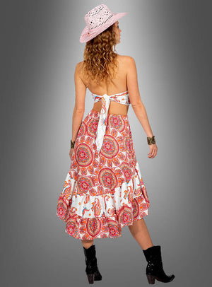 Eine Frau in einem rückenfreien Neckholder-Top und einem gemusterten Rock, ergänzt durch einen rosa Hut und schwarze Stiefel, steht vor einem schlichten grauen Hintergrund mit dem Rücken zur Kamera – ihr „Paisley 2-Teiler mit Rock Festivalstyle“ sorgt für den perfekten Festival-Look eines schicken Boho-Outfits.