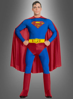 Eine Person, die das „Deluxe Superman-Kostüm für Erwachsene“ trägt – bestehend aus einem blauen Anzug, einem roten Umhang, einem gelben Gürtel und dem ikonischen S-Emblem auf der Brust –, posiert mit den Händen in den Hüften vor einem grauen Hintergrund; dieses offiziell lizenzierte DC-Comics-Design ist ein klassisches Superheldenkostüm.