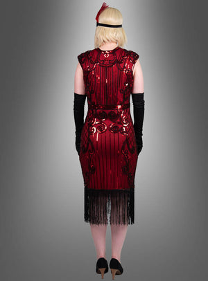 Eine Person trägt das Luxus-Kleid „Kelly“ im Stil der 1920er Jahre – ein auffälliges rotes Paillettenkleid mit schwarzem Fransenbesatz –, kombiniert mit schwarzen Handschuhen, schwarzen High Heels und einem roten, mit Federn verzierten Stirnband. Sie steht vor einem grauen Hintergrund mit Farbverlauf und schaut von der Kamera weg.