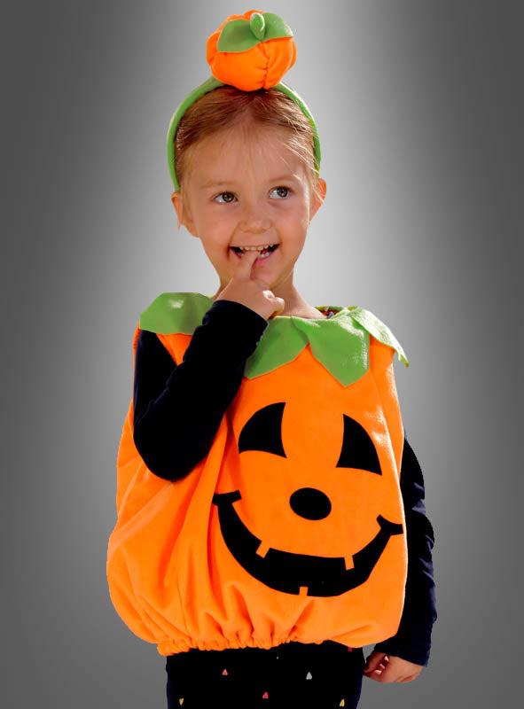 Ein kleines Kind, das das „Deluxe Kürbiskostüm aus Plüsch für Kinder“ trägt – ein Plüschkostüm in Form eines Kürbisses mit einem Jack-o’-Lantern-Gesicht und einer dazu passenden Kürbismütze –, steht vor einem grauen Hintergrund, lächelt und hat einen Finger im Mund.