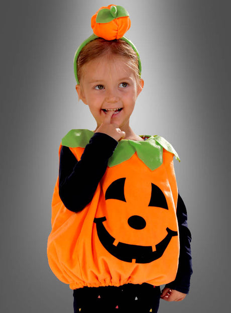 Ein kleines Kind, das das „Deluxe Kürbiskostüm aus Plüsch für Kinder“ trägt – ein Plüschkostüm in Form eines Kürbisses mit einem Jack-o’-Lantern-Gesicht und einer dazu passenden Kürbismütze –, steht vor einem grauen Hintergrund, lächelt und hat einen Finger im Mund.