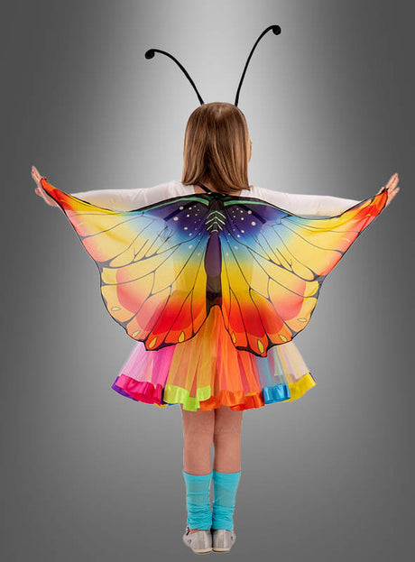 Ein Kind steht mit ausgestreckten Armen vor einem grauen Hintergrund mit Farbverlauf und trägt ein „Schmetterling Flügel“-Kostüm mit leuchtend bunten Flügeln und verspielten Fühlern – ein perfektes Geschenk für Mädchen, die sich gerne verkleiden.