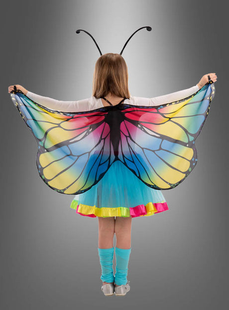 Ein Kind, das das „Schmetterling Flügel“-Kostüm trägt, steht mit ausgestreckten Armen da und präsentiert seine leuchtenden Schmetterlingsflügel vor einem schlichten grauen Hintergrund mit Farbverlauf – ein ideales Geschenk für Mädchen, die fantasievolles Spielen lieben.