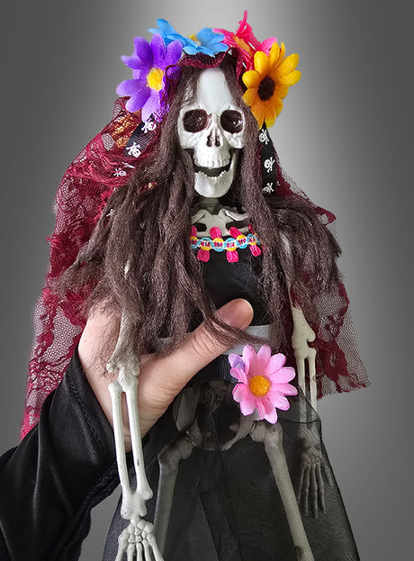 Eine Hand hält die „Tag der Toten“-Voodoo-Puppe „Braut“ (42 cm), eine Skelettpuppe, die mit einem bunten Blumenkopfschmuck, einem Schleier, langem braunem Haar und Blumenaccessoires vor grauem Hintergrund – ideal als „Tag der Toten“-Dekoration für Ihr Zuhause oder als Teil eines Skelett-Brautpaares zur Feier des mexikanischen Halloween.