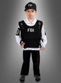 Ein kleines Kind steht vor einem grau abgestuften Hintergrund und trägt ein FBI-Agenten-Kostüm für Kinder, komplett mit Mütze, Weste und weißem Hemd.