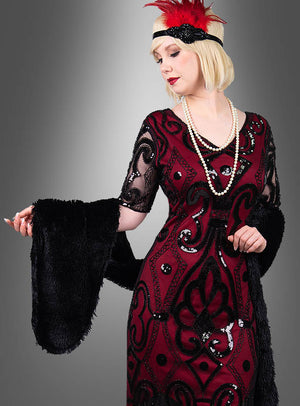 Eine Frau in einem rot-schwarzen Flapper-Kleid mit Pailletten, umhüllt von der „Lange Stola 20er Jahre“ in klassischem Schwarz als elegante Vintage-Stola, ergänzt durch eine lange Perlenkette und ein rotes Stirnband mit Federn – ihr Look wird durch Charleston-Kostüm-Accessoires perfekt abgerundet –, vor einem schlichten grauen Hintergrund.