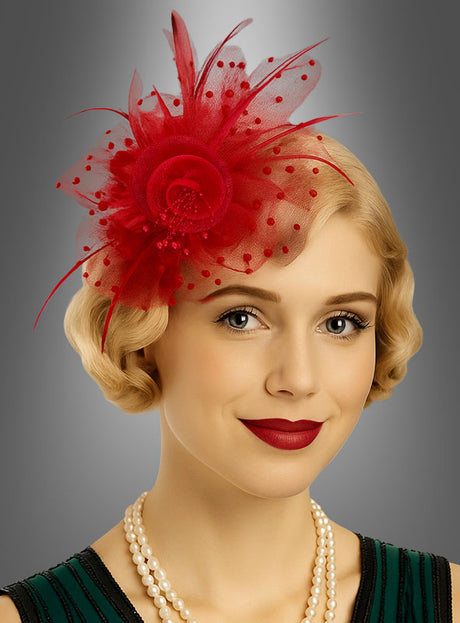 Eine Frau mit kurzen blonden Haaren trägt vor einem grauen Hintergrund mit Farbverlauf einen leuchtend roten „Fascinator Minihut mit Tüll“, ein grünes Kleid, eine Perlenkette und roten Lippenstift – ein elegantes Accessoire, das ihren raffinierten Look mühelos abrundet.