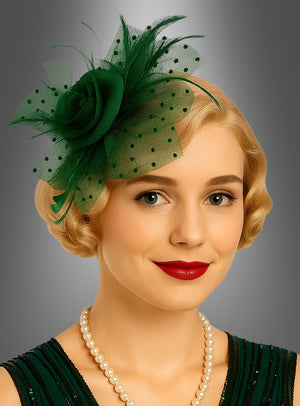 Eine Frau mit kurzen blonden Haaren, rotem Lippenstift und einer Perlenkette trägt einen grünen „Fascinator Minihut mit Tüll“ – ein elegantes Accessoire –, dazu ein passendes grünes Kleid, und posiert vor einem grauen Hintergrund mit Farbverlauf.