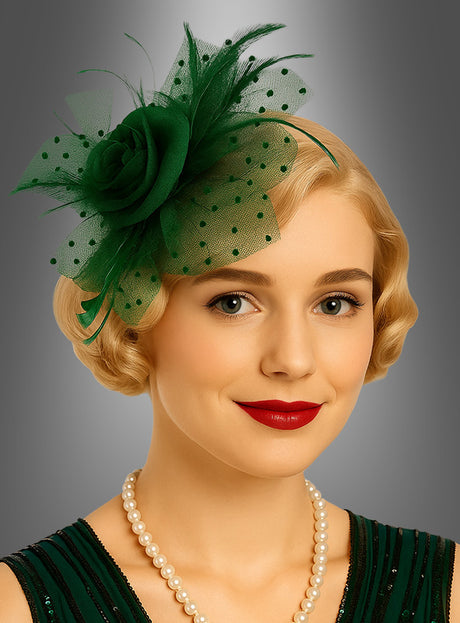 Eine Frau mit kurzen blonden Haaren, rotem Lippenstift und einer Perlenkette trägt einen grünen „Fascinator Minihut mit Tüll“ – ein elegantes Accessoire –, dazu ein passendes grünes Kleid, und posiert vor einem grauen Hintergrund mit Farbverlauf.
