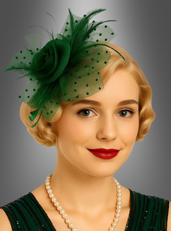 Eine Frau mit kurzen blonden Haaren, rotem Lippenstift und einer Perlenkette trägt einen grünen „Fascinator Minihut mit Tüll“ – ein elegantes Accessoire –, dazu ein passendes grünes Kleid, und posiert vor einem grauen Hintergrund mit Farbverlauf.