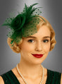 Eine Frau mit kurzen blonden Haaren, rotem Lippenstift und einer Perlenkette trägt einen grünen „Fascinator Minihut mit Tüll“ – ein elegantes Accessoire –, dazu ein passendes grünes Kleid, und posiert vor einem grauen Hintergrund mit Farbverlauf.
