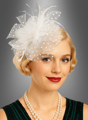 Eine Frau mit kurzen blonden Haaren trägt einen weißen, mit Polka-Dots verzierten Fascinator-Minihut mit Tüll als elegantes Accessoire, knallroten Lippenstift und eine zweireihige Perlenkette und posiert vor einem grauen Hintergrund.