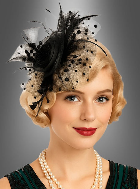 Eine Frau mit kurzen blonden Haaren trägt ein elegantes Accessoire: den „Fascinator Minihut mit Tüll“, einen schwarz-beigen Kopfschmuck, der mit Tüll und Federn verziert ist und wie ein Minihut gestaltet ist. Sie kombiniert ihn mit einem grünen Kleid und mehrreihigen Perlenketten und posiert vor einem grauen Hintergrund mit Farbverlauf.
