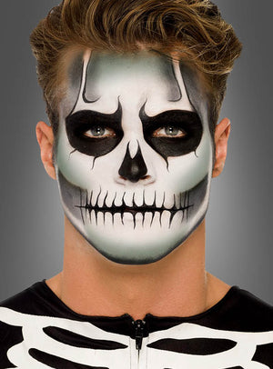 Eine Person mit hellbraunen Haaren trägt das „Leuchtendes Skelett“-Make-up-Set und ein Skelettkostüm und blickt vor einem grauen Hintergrund direkt in die Kamera.