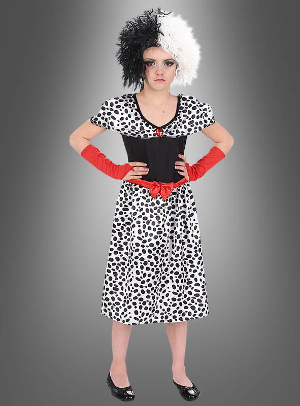 Ein Mädchen, das das Cruella-Dalmatiner-Kostüm für Mädchen trägt – ein schwarz-weiß geflecktes Kleid mit einem roten Gürtel, dazu eine passende schwarz-weiße Perücke und rote Handschuhe –, steht vor einem grauen Hintergrund.