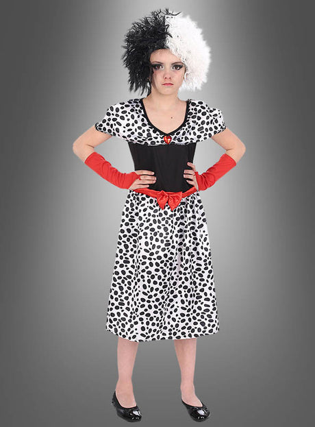 Ein Mädchen, das das Cruella-Dalmatiner-Kostüm für Mädchen trägt – ein schwarz-weiß geflecktes Kleid mit einem roten Gürtel, dazu eine passende schwarz-weiße Perücke und rote Handschuhe –, steht vor einem grauen Hintergrund.