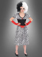 Ein Mädchen, das das Cruella-Dalmatiner-Kostüm für Mädchen trägt – ein schwarz-weiß geflecktes Kleid mit einem roten Gürtel, dazu eine passende schwarz-weiße Perücke und rote Handschuhe –, steht vor einem grauen Hintergrund.