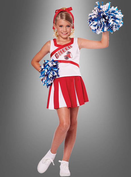 Ein junges Mädchen, das das Cheerleader-Kostüm für Kinder mit Pompons trägt und blaue und weiße Pompons in den Händen hält, steht lächelnd vor einem grauen Hintergrund.