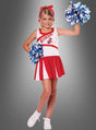 Ein junges Mädchen, das das Cheerleader-Kostüm für Kinder mit Pompons trägt und blaue und weiße Pompons in den Händen hält, steht lächelnd vor einem grauen Hintergrund.