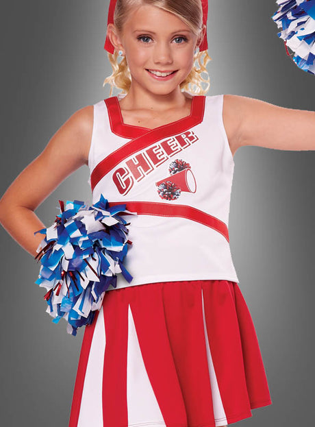 Ein junges Mädchen im Cheerleader-Kostüm für Kinder mit Pompons in Rot und Weiß, das blaue und weiße Pompons in den Händen hält und vor einem grauen Hintergrund lächelt.