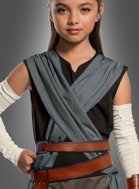 Ein Kind, das das Rey-Kostüm für Kinder aus „Star Wars VIII“ trägt – bestehend aus einer dunklen Tunika, grauen Wickeltüchern, weißen Armstulpen und einem braunen Gürtel –, steht vor einem einfarbigen Hintergrund.