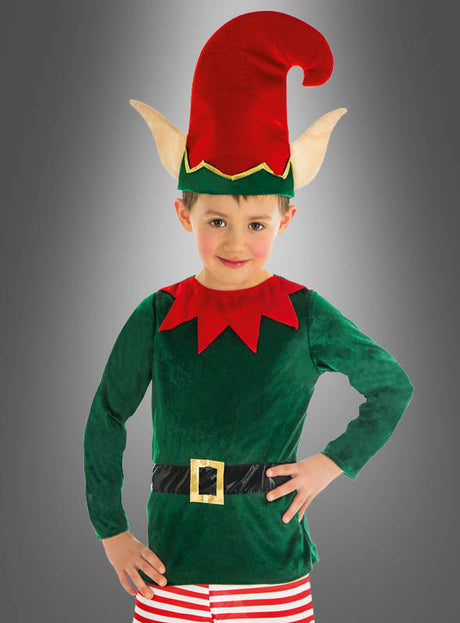 Ein Kind, das das „Weihnachtselfe“-Kinderkostüm trägt – ein grünes Elfenkostüm aus Samt mit rotem Kragen, schwarzem Gürtel, gestreifter Hose und einer roten Mütze mit großen, spitzen Ohren –, steht vor einem grauen Hintergrund mit Farbverlauf; perfekt als Weihnachtskostüm für Kinder oder Fasching-Kostüm für Kinder für festliche Anlässe.