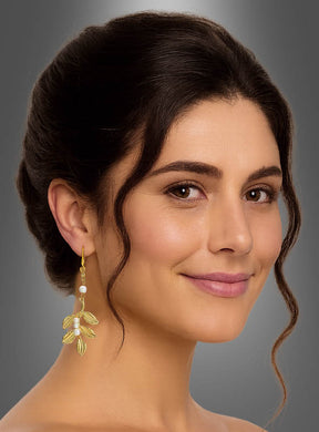 Eine Frau mit dunklem, hochgestecktem Haar, die die eleganten Ohrringe „Lorbeerblatt mit Perlen“ in Gelbgold trägt, lächelt sanft vor einem grauen Hintergrund.