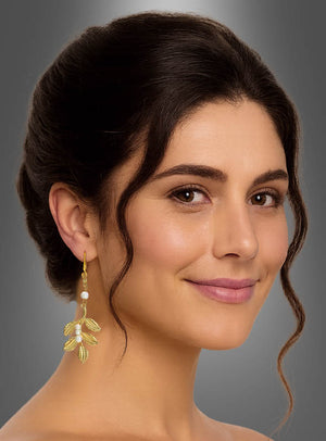 Eine Frau mit dunklem, hochgestecktem Haar, die die eleganten Ohrringe „Lorbeerblatt mit Perlen“ in Gelbgold trägt, lächelt sanft vor einem grauen Hintergrund.