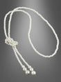 Die „Deluxe Perlenkette Modeschmuck creme“ ist eine lange, einsträngige Kette aus weißen Perlen, die nahe einem Ende geknotet ist und zwei lose Enden aufweist, die jeweils mit größeren runden Perlen abgeschlossen sind; sie wird vor einem grauen Hintergrund mit Farbverlauf präsentiert.