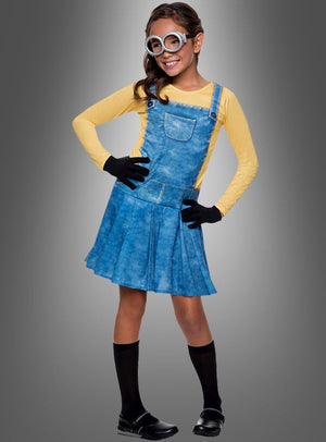 Ein Kind im Minion-Mädchenkostüm, bestehend aus einem gelben Shirt, einem blauen Latzkleid, schwarzen Handschuhen, schwarzen Kniestrümpfen, schwarzen Schuhen und einer großen runden Schutzbrille, steht vor einem grauen Hintergrund.