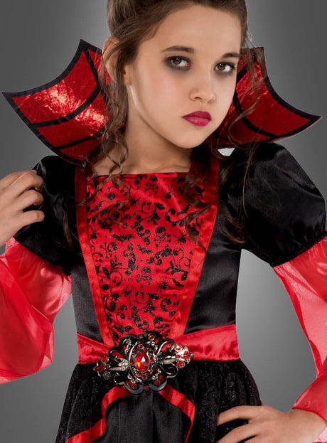 Ein Mädchen in einem dramatischen Vampirgräfin-Kostüm für Mädchen mit auffälligen schwarzen und roten Details, einem markanten Kragen, dunklem Augen-Make-up und glänzendem Lippenstift, das vor einem grauen Hintergrund posiert – das ideale Halloween-Kostüm für Mädchen für einen gruseligen Abend.