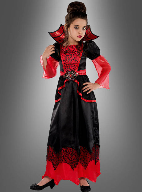 Ein Mädchen, das das auffällige „Vampirgräfin“-Kostüm für Mädchen trägt – ein elegantes rot-schwarzes Vampir-Kleid mit hohem Kragen –, steht vor einem grauen Hintergrund, was dieses Halloween-Kostüm zu einem echten Highlight für Mädchen macht.