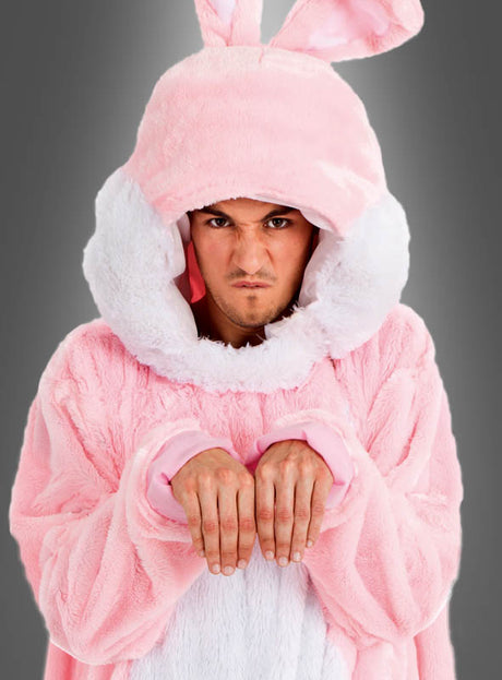 Eine Person, die das Kostüm „Rosa Plüsch Hase für Damen oder Herren“ mit Kapuze und Hasenohren trägt, hält die Hände wie Pfoten vor sich und blickt mit grimmigem Gesichtsausdruck vor einem grauen Hintergrund in die Kamera.
