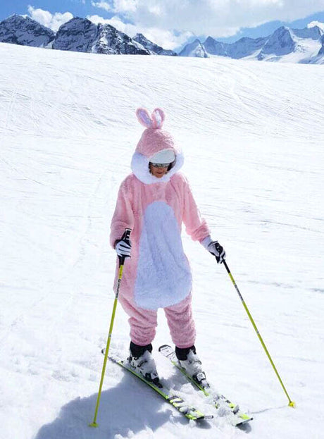 Eine Person, die den „Rosa Plüsch Hase für Damen oder Herren“ trägt, fährt unter strahlend blauem Himmel einen verschneiten Berghang hinunter, umgeben von schneebedeckten Gipfeln.