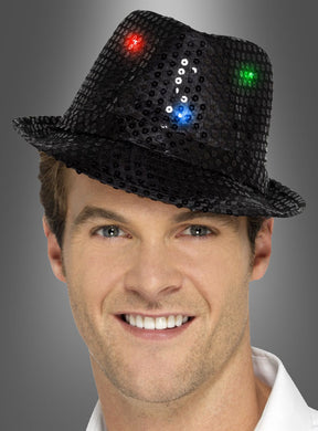 Eine lächelnde Person, die einen schwarzen Trilby-Hut mit LED trägt, der mit roten, blauen und grünen Lichtern verziert ist, vor einem grauen Hintergrund.